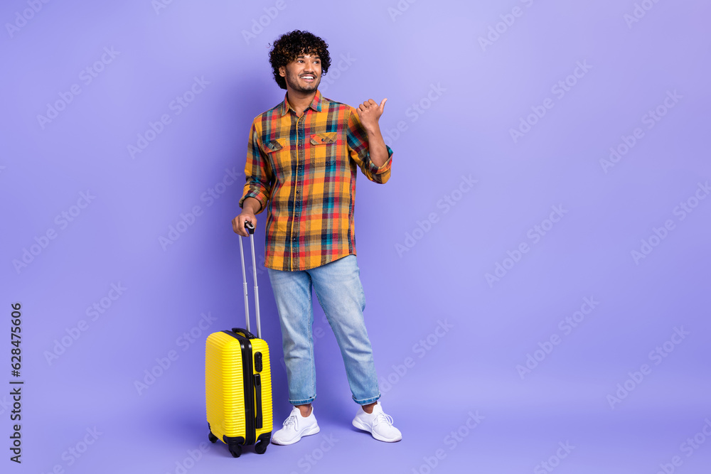 Full body photo of young guy wear trendy jeans direct finger mockup schedule airport carry suitcase isolated on violet color background Full body photo of young guy wear trendy jeans direct finger mockup schedule airport carry suitcase isolated on violet color background
