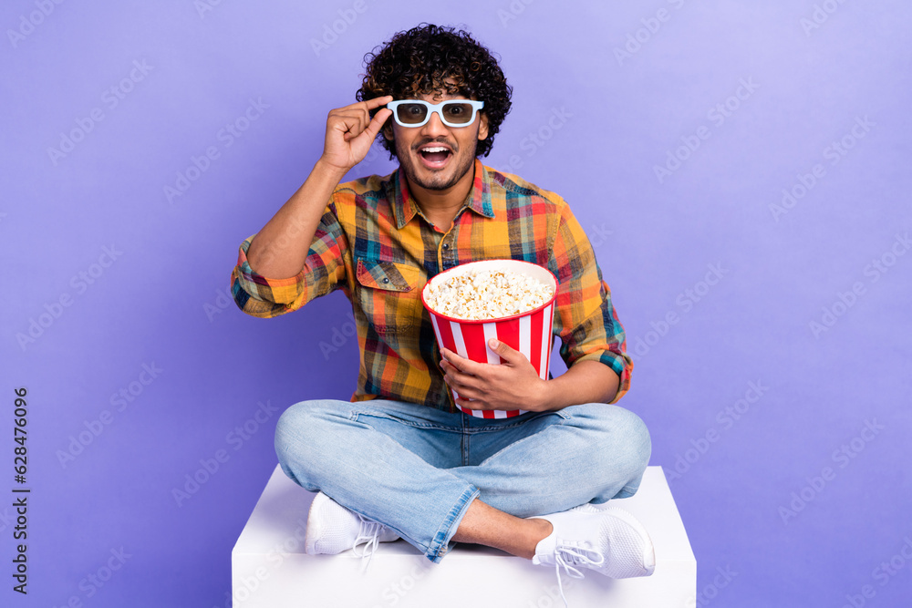 Full size portrait of astonished person sit podium hold popcorn bucket hand touch 3d glasses isolated on purple color background