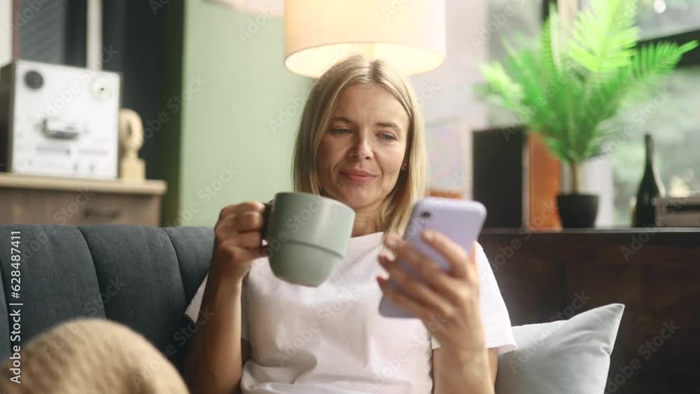 Portrait of charming blond mature woman with cup of hot coffee or tea hold smartphone scrolling watching social media indoors Happy female texting on her phone enjoying leisure time at home Portrait of charming blond mature woman with cup of hot coffee or tea hold smartphone scrolling watching social media indoors Happy female texting on her phone enjoying leisure time at home