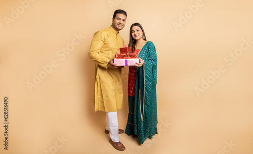 Happy young indian couple wearing traditional cloths holding red gift boxes celebrating of diwali festival isolated on beige background. Copy space