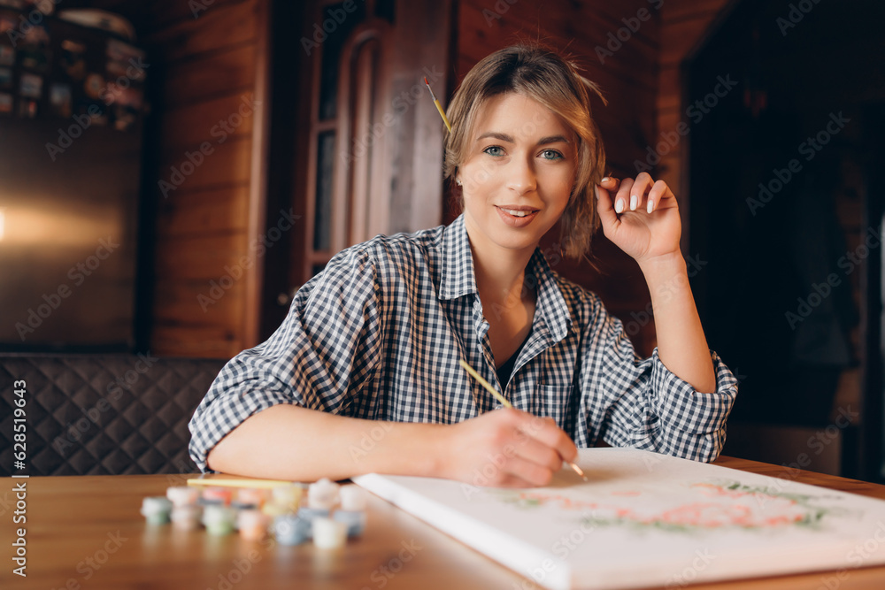 Fototapeta premium A beautiful young woman artist is working in her studio, holding a brush and smiling. She is looking at the camera. Inspiration, creative skills and artistic concept