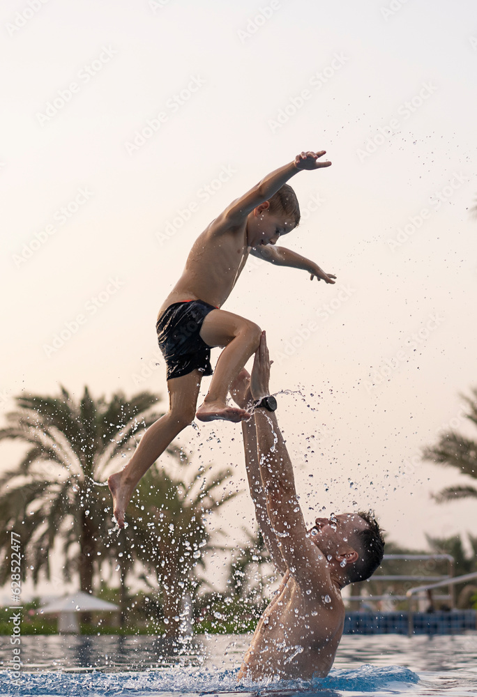 Dad and son are playing in the swimming pool. The father tosses the son up. 