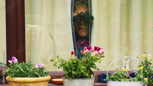 Closeup from the outside of a senior man in a room looking out through a gap in the curtains with a pair of binoculars, at something or someone in the distance.