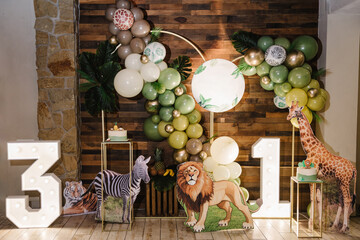 Arch decorated green, golden balloons, neon numbers, paper leaves. Birthday cake for 1 and 3 year old girl or boy on photo wall. Children's photo zone. Reception at birthday party. Decor in style zoo