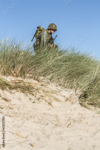 Historical reeneactor dressed as World War II-era infantry soldier views the site while kneeling among the tall grass 