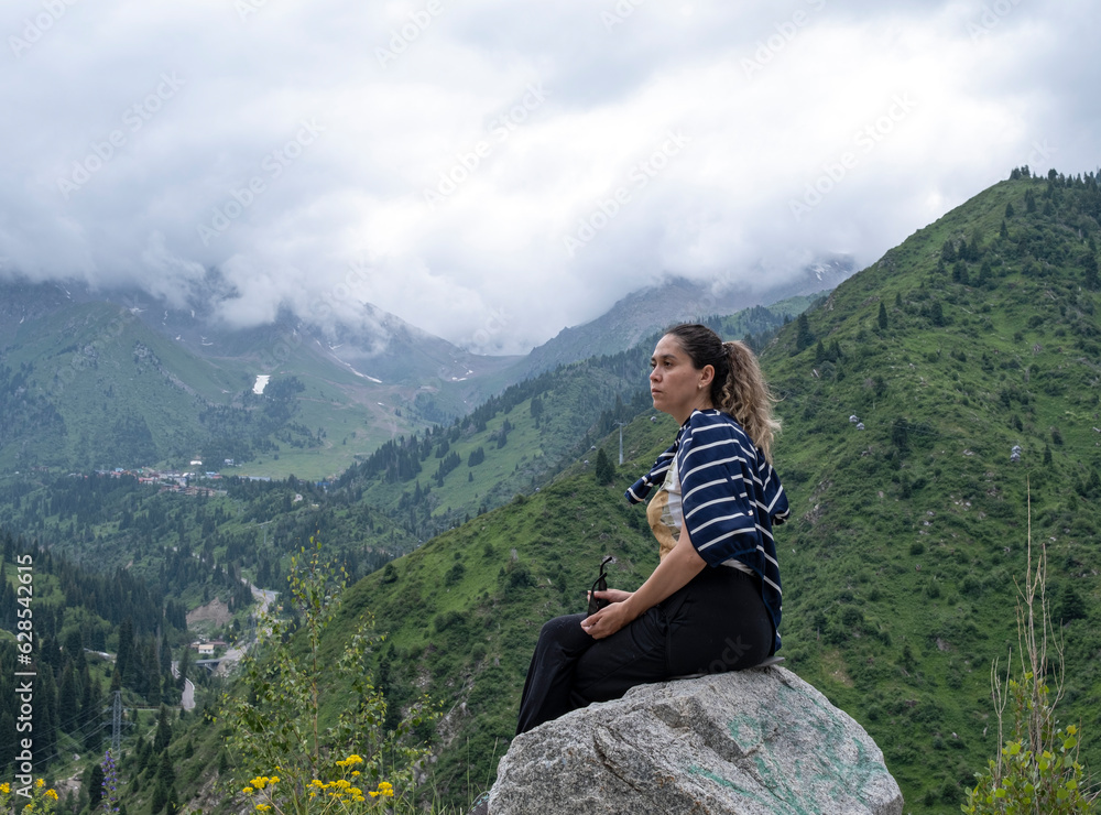 A young woman sits on a rock and looks into the distance. A woman is resting and admiring the view of the mountain valley. There are mountains and clouds in the background.