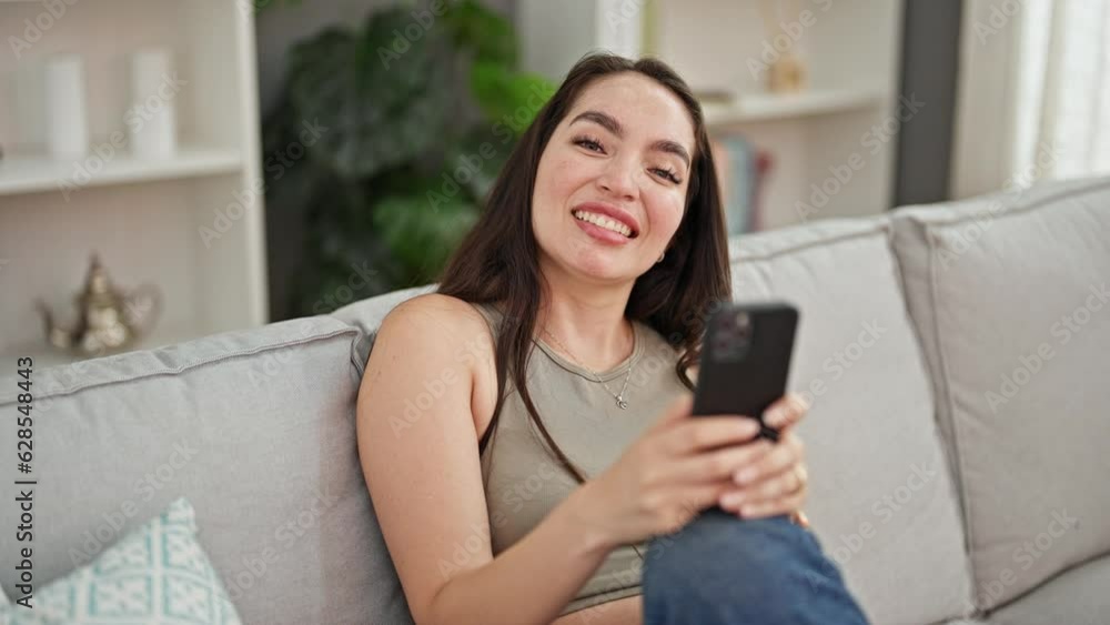 Young beautiful hispanic woman using smartphone sitting on sofa at home Young beautiful hispanic woman using smartphone sitting on sofa at home