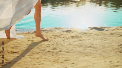 women's legs feet close-up runs walks along beach river banks, stepping barefoot on yellow sand. Body part happy Woman enjoys nature free life water sun light. Girl go in white long silk dress train photo