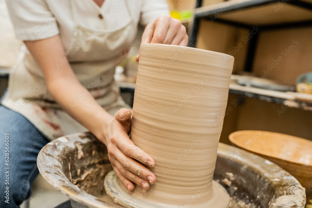 Cropped view of blurred craftswoman in apron creating clay vase on pottery wheel in workshop Cropped view of blurred craftswoman in apron creating clay vase on pottery wheel in workshop