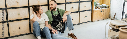 Smiling couple of sculptors sitting on floor near cupboard in ceramic workshop, banner photo