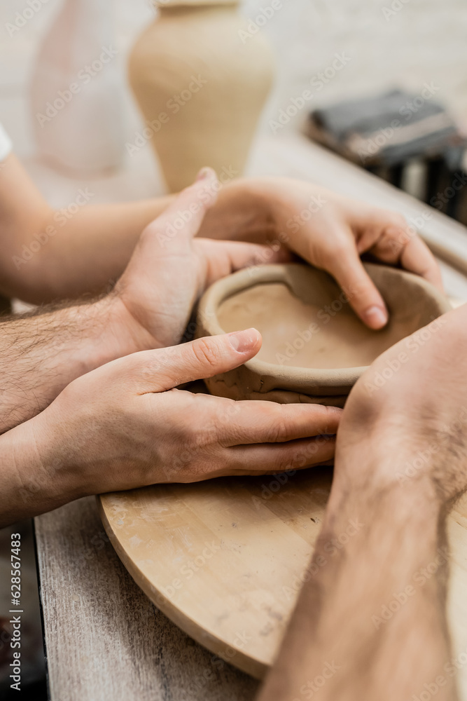 Cropped view of couple of artisan shaping clay bowl near table in ceramic workshop Cropped view of couple of artisan shaping clay bowl near table in ceramic workshop