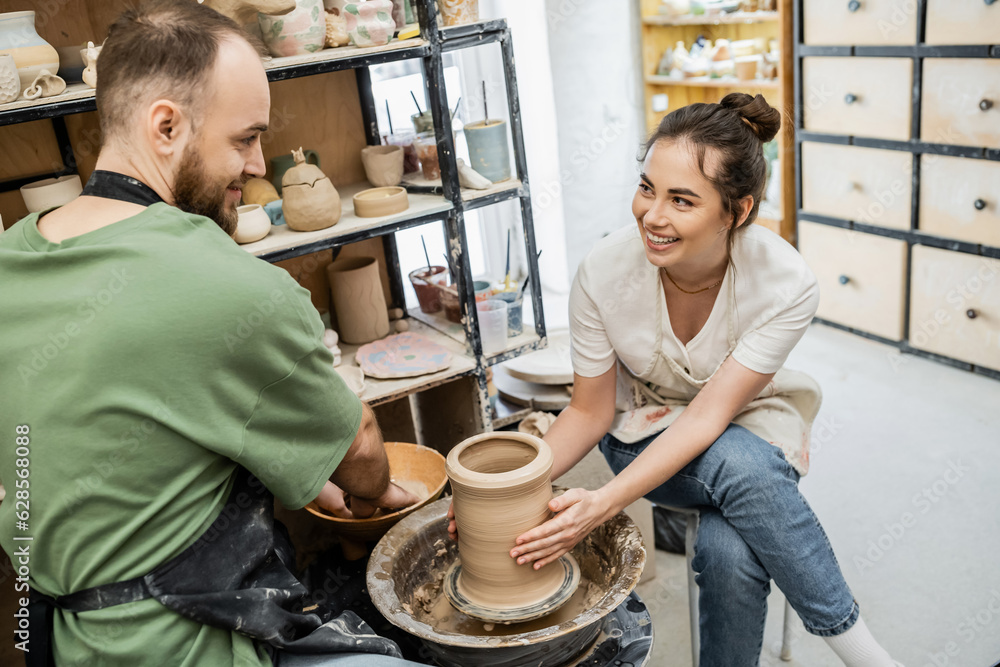 Smiling craftswoman shaping clay vase on pottery wheel near boyfriend and bowl with water in studio