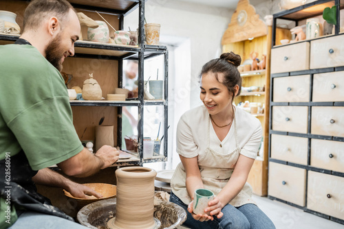 Smiling craftswoman holding cup while boyfriend making clay vase on pottery wheel in studio photo