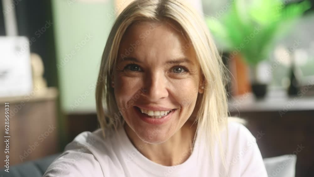 Camera view of smiling middle aged woman looking at the camera and have a video call conference meeting with family or friends at home Happy blond female has pleasant conversation indoors Camera view of smiling middle aged woman looking at the camera and have a video call conference meeting with family or friends at home Happy blond female has pleasant conversation indoors