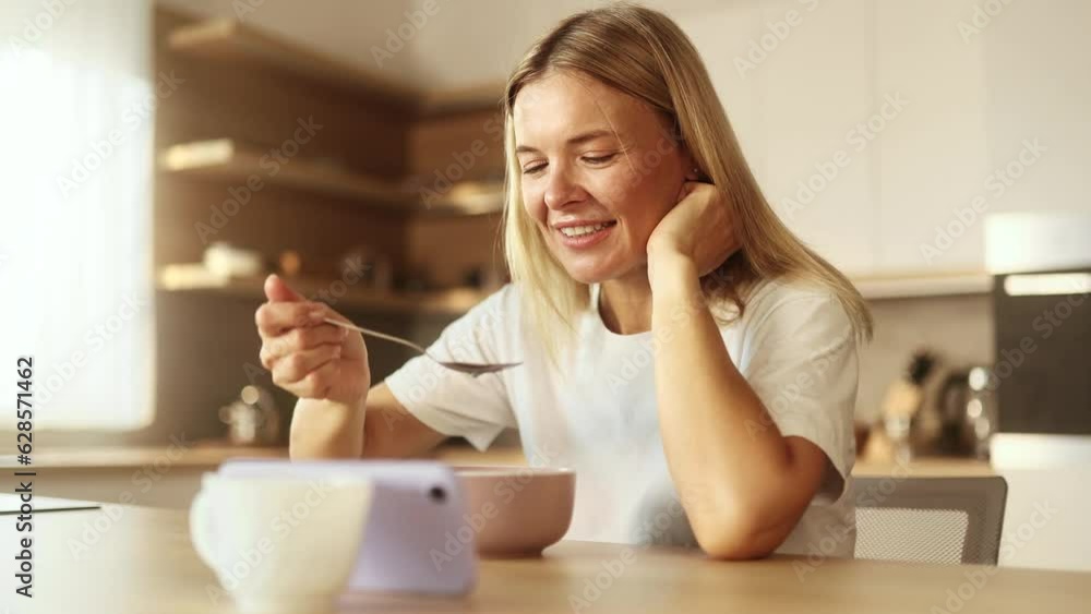 Pretty blond mature woman enjoying beautiful morning with tasty breakfast laughing while watching online funny videos entertainment programs in social media on smartphone in sunny kitchen at home Pretty blond mature woman enjoying beautiful morning with tasty breakfast laughing while watching online funny videos entertainment programs in social media on smartphone in sunny kitchen at home