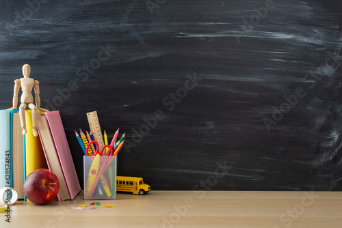 School supplies layout. Side view photo of desk setup with pencils organizer, ruler, books, red apple, mannequin body, and more on chalkboard background. Great for educational content or advertising