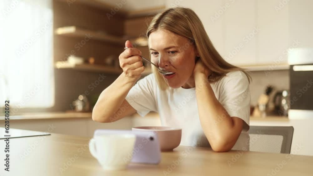 Pretty blond mature woman enjoying beautiful morning with tasty breakfast laughing while watching online funny videos entertainment programs in social media on smartphone in sunny kitchen at home Pretty blond mature woman enjoying beautiful morning with tasty breakfast laughing while watching online funny videos entertainment programs in social media on smartphone in sunny kitchen at home