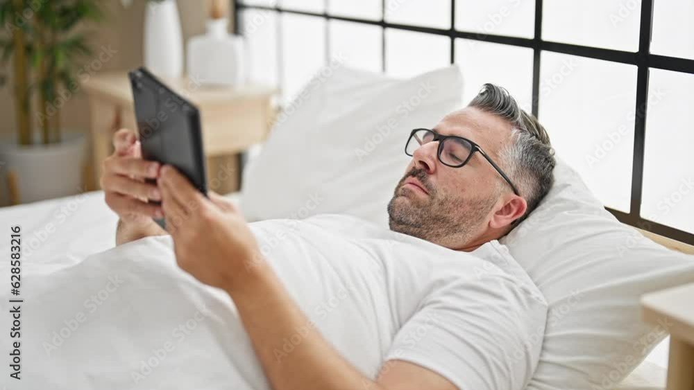 Grey-haired man using touchpad taking glasses off to sleep at bedroom Grey-haired man using touchpad taking glasses off to sleep at bedroom