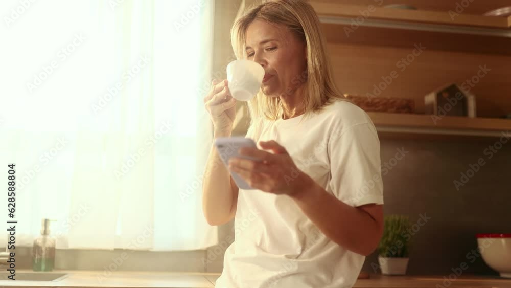 Pretty mature woman enjoying beautiful morning with cup of coffee using smartphone scrolling social media at sunny kitchen Charming blond female relaxing and chatting on mobile phone at home Pretty mature woman enjoying beautiful morning with cup of coffee using smartphone scrolling social media at sunny kitchen Charming blond female relaxing and chatting on mobile phone at home