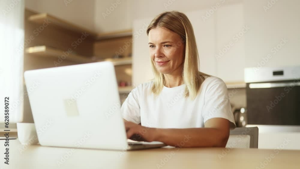 Portrait of blond mature woman working online typing browsing products in internet store reading news checking email or social media profile on laptop computer at light home indoors Portrait of blond mature woman working online typing browsing products in internet store reading news checking email or social media profile on laptop computer at light home indoors