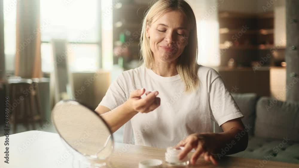 Portrait of pretty mature blond woman looking at mirror reflection and applying cream on hands moisturising skin at home Concept of skincare cosmetology natural beauty cosmetics Portrait of pretty mature blond woman looking at mirror reflection and applying cream on hands moisturising skin at home Concept of skincare cosmetology natural beauty cosmetics