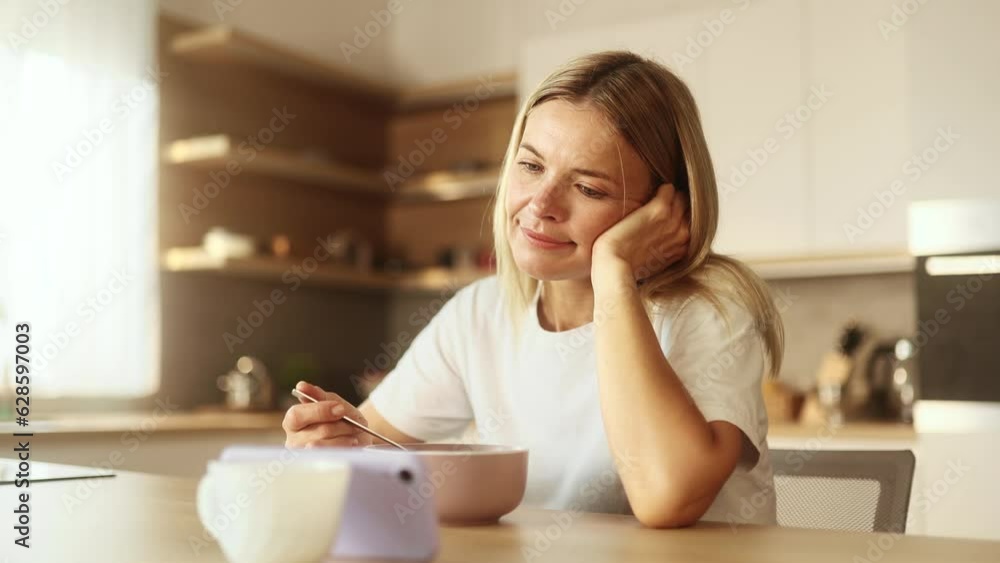 Boring exhausted mature woman watching online videos entertainment programs or news in social media on smartphone gadget while having breakfast in the morning at home Boring exhausted mature woman watching online videos entertainment programs or news in social media on smartphone gadget while having breakfast in the morning at home