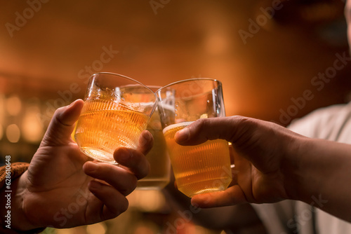 Close-up of a group of three friends standing at the bar talking and toast with glasses of beer and a cocktail in a club bar