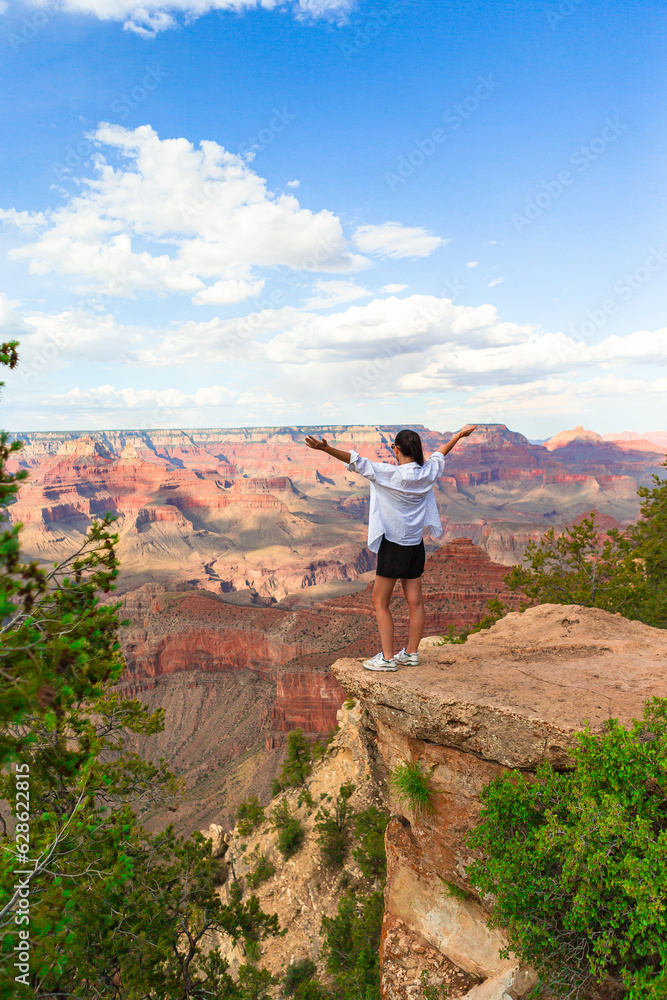Fototapeta premium Happy young woman on a steep cliff taking in the amazing view over famous Grand Canyon at sunset, Grand Canyon National Park, Arizona, USA