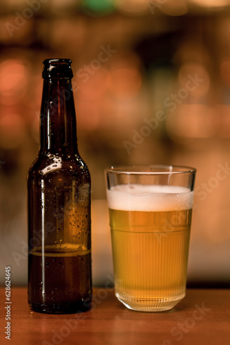 Close up of a bottle of beer standing next to a glass of beer on a bar counter in a club bar on a blurred background