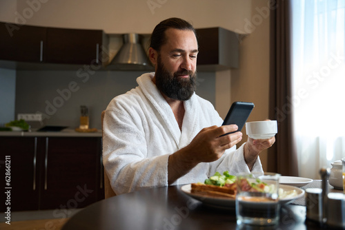 Male in a bathrobe sits at a table with a phone