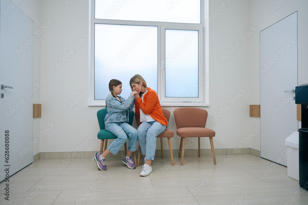 Girl with her mother are sitting on armchairs in hospital corridor