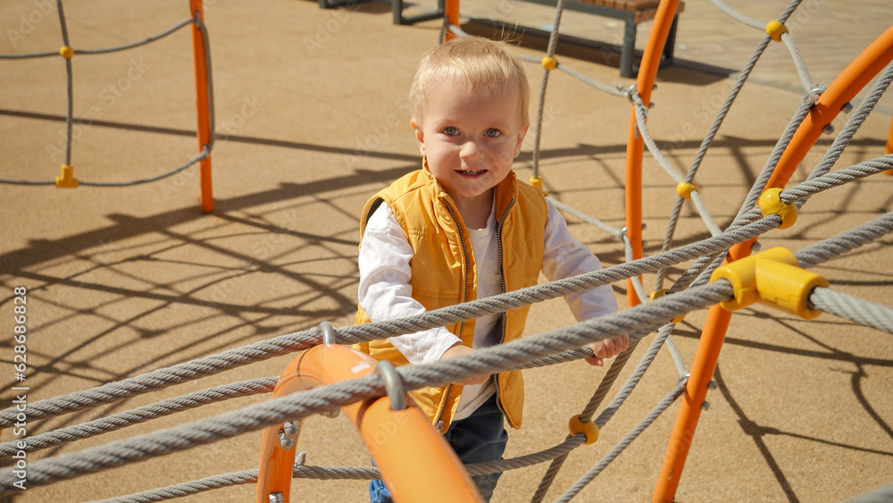 Portrait of cute cheerful baby boy climbing up on the rope ladder at playground. Children playing outdoor, kids outside, summer holiday and vacation. Portrait of cute cheerful baby boy climbing up on the rope ladder at playground. Children playing outdoor, kids outside, summer holiday and vacation.