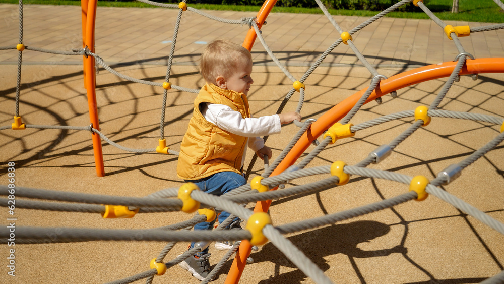 Little baby boy climbing up the spider web on playground. Children playing outdoor, kids outside, summer holiday and vacation.