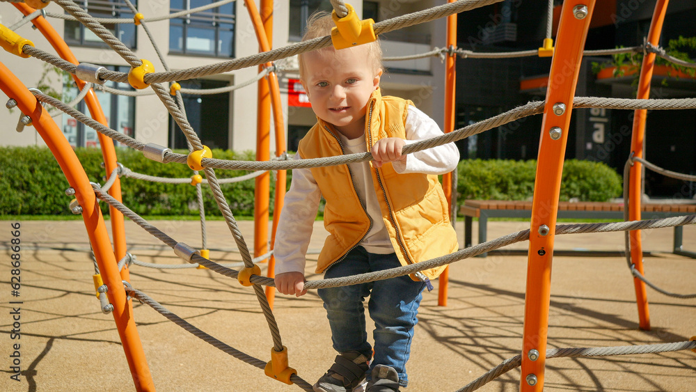 Cute smiling toddler boy climbing up high on the rope ladder at playground