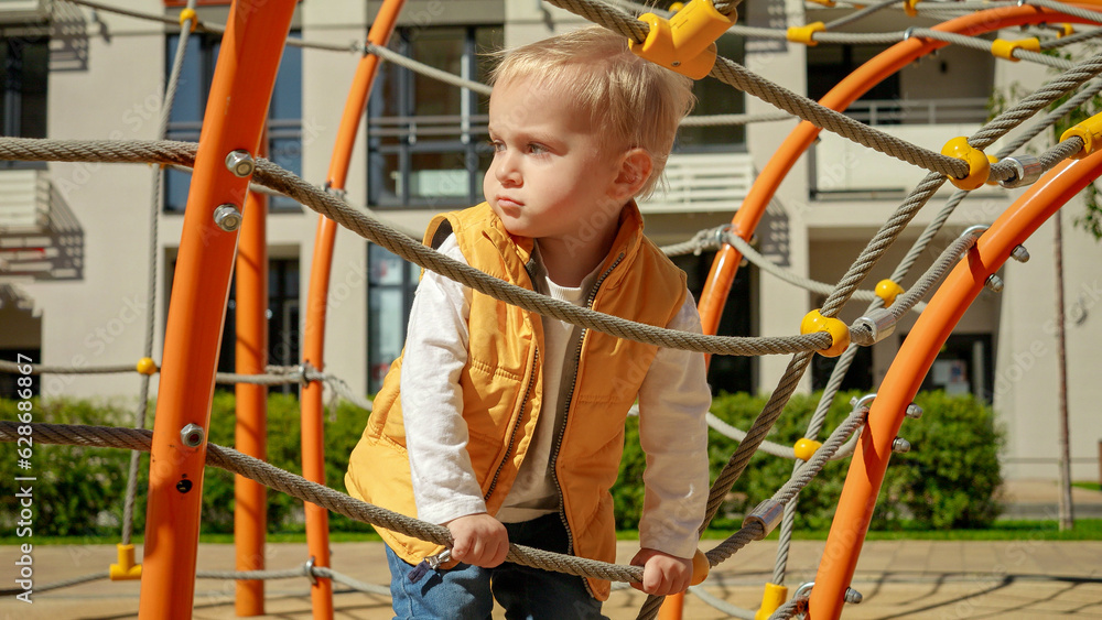 Cute baby boy smiling and having fun climbing up the ropes and nets on playground. Children playing outdoor, kids outside, summer holiday and vacation.
