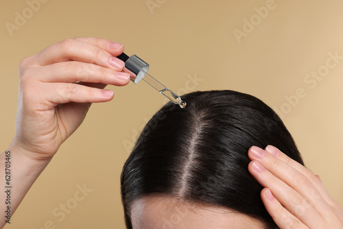 Woman applying essential oil onto hair roots on beige background, closeup