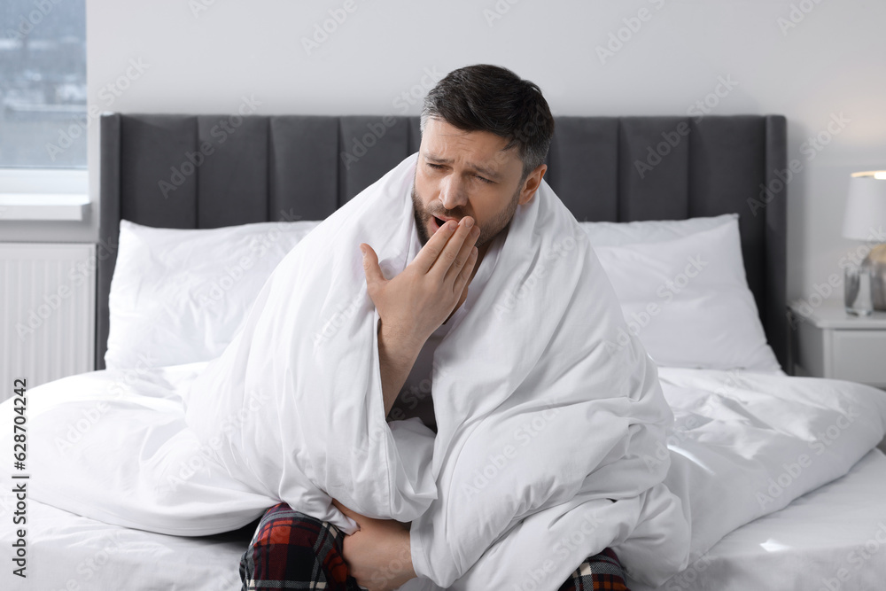 Sleepy man wrapped in blanket yawning on soft bed at home Sleepy man wrapped in blanket yawning on soft bed at home