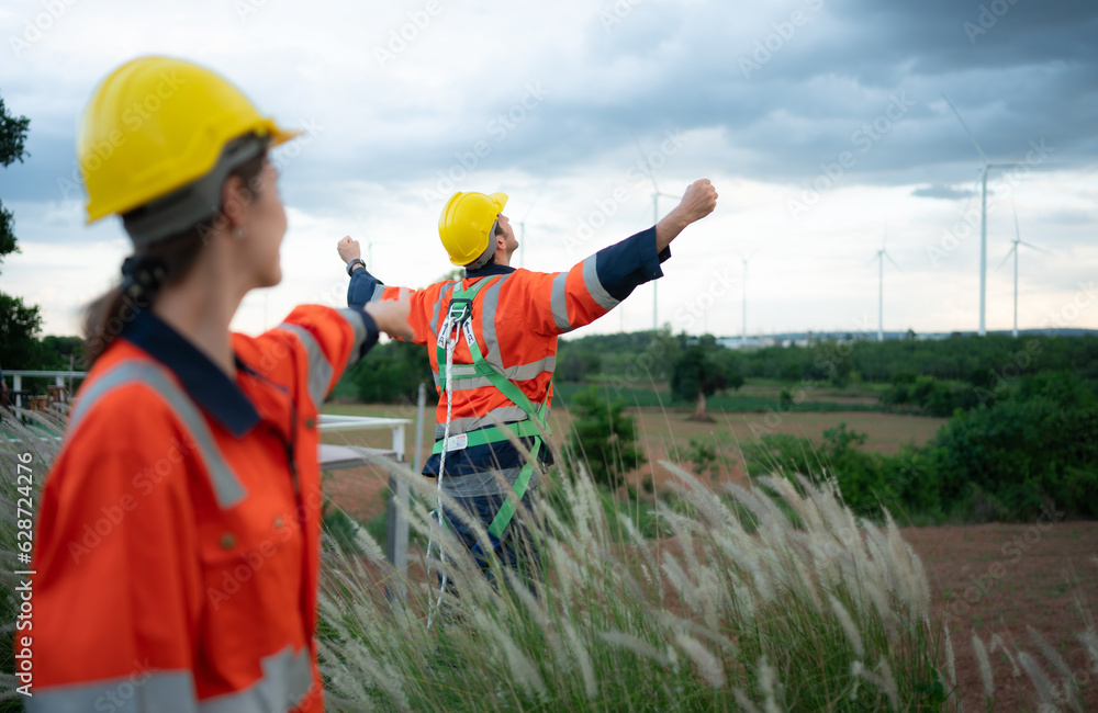 Young female engineer working in a wind turbine field and young man engineer wear a safety vest raise both hands to relax after finishing the wind turbine inspection mission Young female engineer working in a wind turbine field and young man engineer wear a safety vest raise both hands to relax after finishing the wind turbine inspection mission