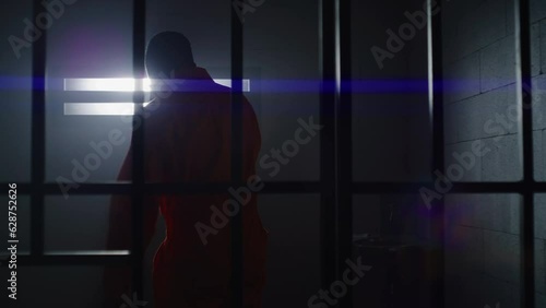 Depressed African American man in orange uniform stands from jail bed, walks at prison cell and looks at barred window. Sad prisoner serves imprisonment term. Murderer in gloomy correctional facility.