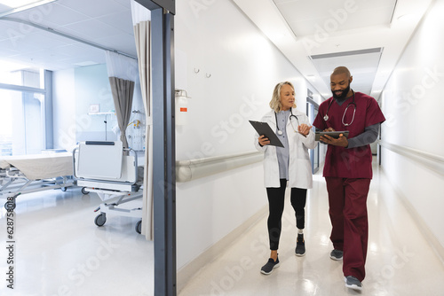 Happy diverse female and male doctors using tablet, talking and walking in corridor at hospital