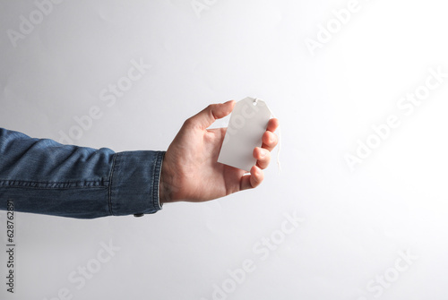 Man's hand in denim shirt holding white blank price tag on gray background. Sale concept