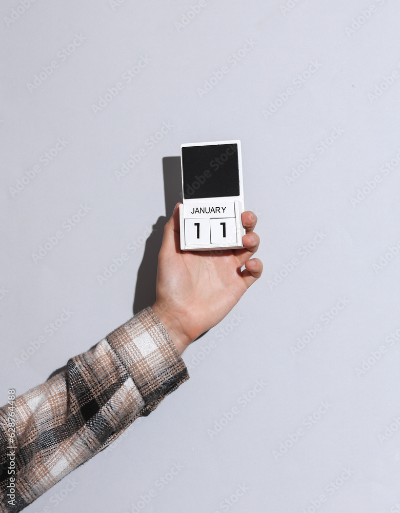 Man's hand in shirt holds wooden block calendar organizer with date january 11 on gray background with shadow Man's hand in shirt holds wooden block calendar organizer with date january 11 on gray background with shadow