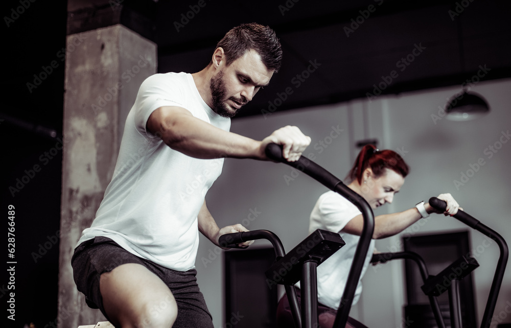 Athletic man and woman doing intense workout together on air bike in modern gym. Healthy lifestyle Athletic man and woman doing intense workout together on air bike in modern gym. Healthy lifestyle