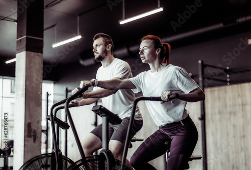 Athletic man and woman doing intense workout together by pedaling air bike in modern gym. Healthy lifestyle