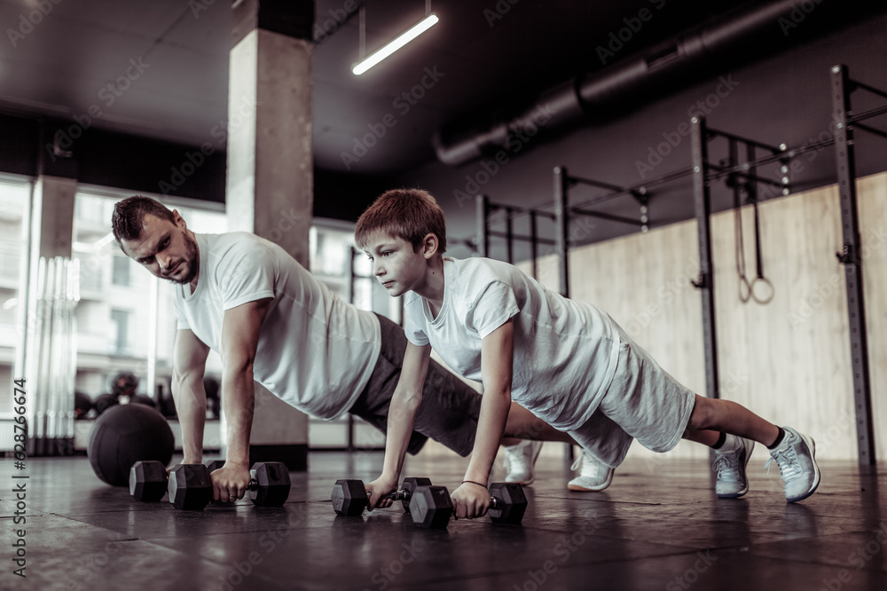 Healthy family concept. Father trainer and teenager son push ups with dumbbells in gym. Fitness, sports, active lifestyle Healthy family concept. Father trainer and teenager son push ups with dumbbells in gym. Fitness, sports, active lifestyle