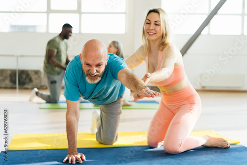 Woman helps mature man to stand in bird dog pose in gym