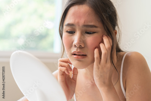 Asian young woman looking face skin in the mirror have a red rash on her face from cosmetic allergy photo