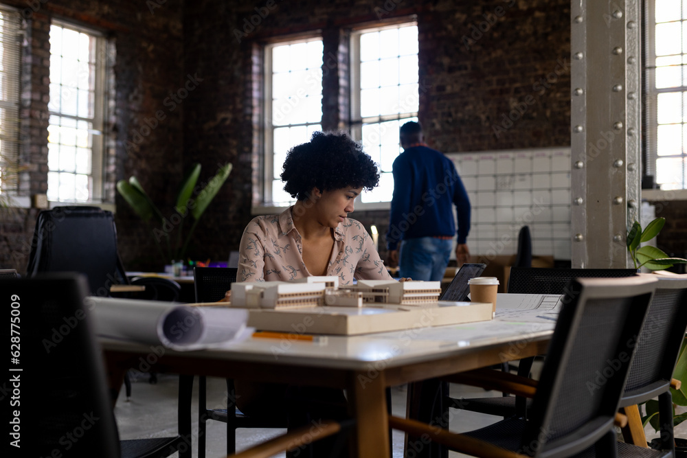 Biracial female architect sitting at desk using tablet and looking at building model in office Biracial female architect sitting at desk using tablet and looking at building model in office