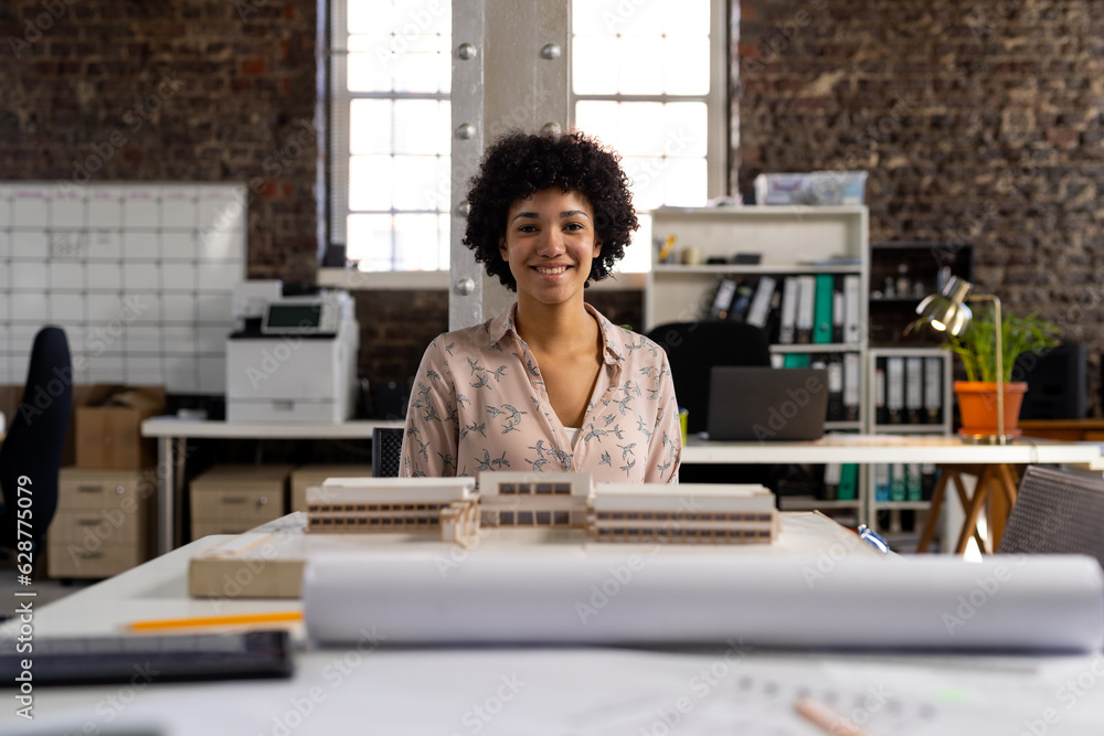 Portrait of happy biracial female architect sitting at desk with building model at office Portrait of happy biracial female architect sitting at desk with building model at office