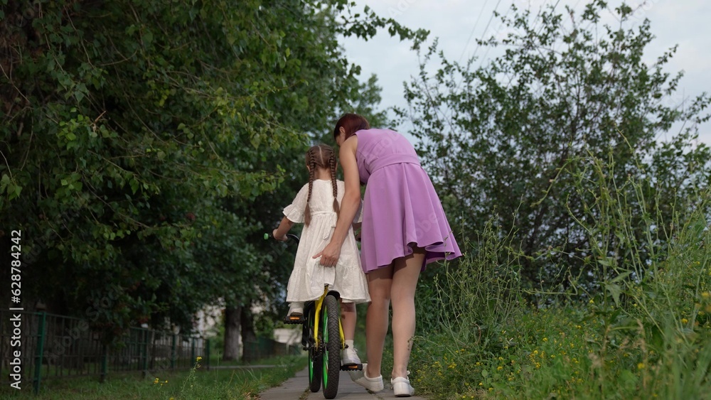 Attentive mother helps cute daughter to ride bicycle in city park backside view